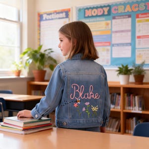 May include: A light blue denim jacket featuring custom embroidery on the back. The design includes the name Blake in pink script lettering above a colorful garden scene with three embroidered flowers and a small bee with a dotted flight path.