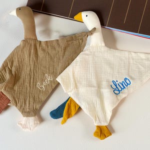 Könnte beinhalten: Zwei gänseförmige Baby-Kuscheltücher. Eines ist beige mit dem Namen "Beck" in Weiß gestickt, das andere ist weiß mit "Lino" in Blau. Beide haben Flügel aus strukturiertem Stoff und farbige Akzente an Füßen und Schwanz.