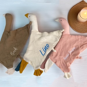 Könnte beinhalten: Drei Baby-Kuscheltücher in Form von Gänsen in Braun, Creme und Rosa. Jedes hat einen gestickten Namen: "Beck", "Lino" und "Annie Jausen". Eine brennende Kerze steht auf einem gewebten Untersetzer.