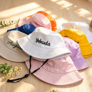 Könnte beinhalten: Eine Auswahl an bunten Fischerhüten in Weiß, Rosa, Gelb und Blau. Einige Hüte haben gestickte Schriftzüge, darunter "Yehuda" und "August". Die Hüte sind auf einer hellen Holzoberfläche angeordnet.