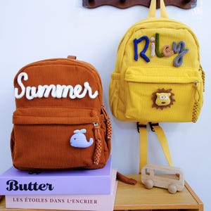 Puede incluir: Una mochila de pana color óxido con el texto blanco Summer y un parche de ballena azul, y una mochila de pana amarilla con el texto multicolor Riley y un parche de león. Ambas mochilas tienen tiradores de cremallera trenzados.