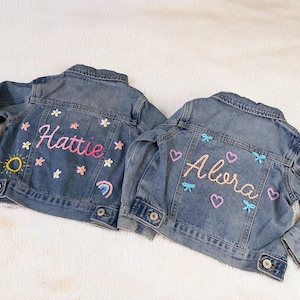 May include: Two blue denim jackets with embroidered names. One jacket says "Hattie" in pink, with flower and rainbow details. The other says "Alora" in pink, with heart and bow details.