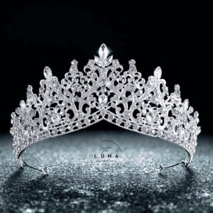 Może przedstawiać: Srebrna tiara z misternym filigranem i błyszczącymi, przezroczystymi kryształami. Korona ma centralny kryształ w kształcie łzy i zakrzywioną podstawę. Widoczne jest logo Luna Design Studio.