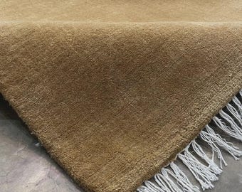 May include: A rectangular area rug in a solid tan colour with a dense, short-pile texture. The rug is folded over to reveal the underside and features a row of long, white, twisted fringe tassels along the straight edge of the fabric.
