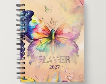 May include: A spiral-bound planner with a watercolor-style cover featuring a large, multi-colored butterfly in shades of blue, pink, yellow, and purple against a soft peach background. The text PLANNER 2027 is printed in dark purple letters.