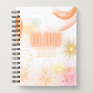 May include: A spiral-bound notebook with a white cover featuring the word "GLOW" in orange, with "2026" below. The design includes orange and yellow abstract shapes and a gold spiral binding.