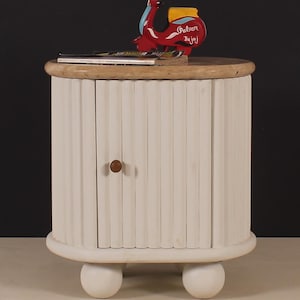 May include: A white, round bedside cabinet with a light wood top and two spherical feet. The cabinet has a ribbed front and a small, round wooden knob. A red scooter figurine with the text "Palian Bajaj" sits on top, along with a stack of magazines.