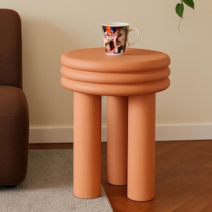 Puede incluir: Una mesa auxiliar de color melocotón con tres patas cilíndricas y una parte superior redonda. Una taza con un diseño abstracto colorido está sobre la mesa. Una obra de arte abstracta enmarcada cuelga de la pared detrás de la mesa.