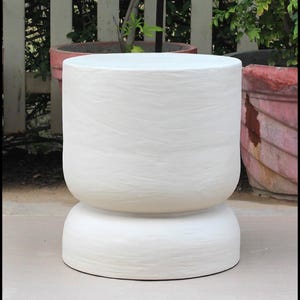 May include: A white, cylindrical side table with a textured surface. The table has a rounded base and a wider top, creating a unique, modern design. The table is approximately 18 inches tall.