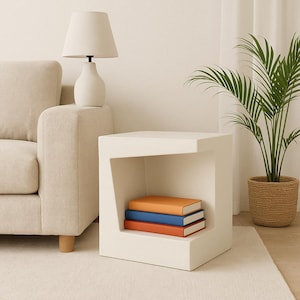 May include: A contemporary white side table with a distinctive cut-out feature. Three books in orange, blue, and red rest inside. A cream lamp sits on a beige sofa, and a potted plant is also visible.
