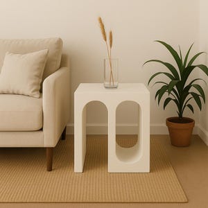 May include: A white side table with arched cutouts, beside a beige sofa and a potted plant. A clear glass vase with wheat stalks sits on the table. The table is on a woven beige rug.