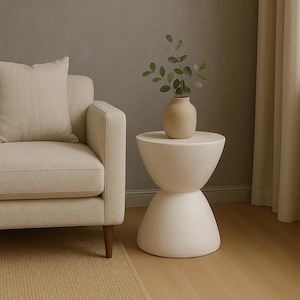 May include: A cream-colored hourglass-shaped side table with a round top and base. A small beige vase with green leafy branches sits on top. The table is next to a beige sofa and a beige rug.