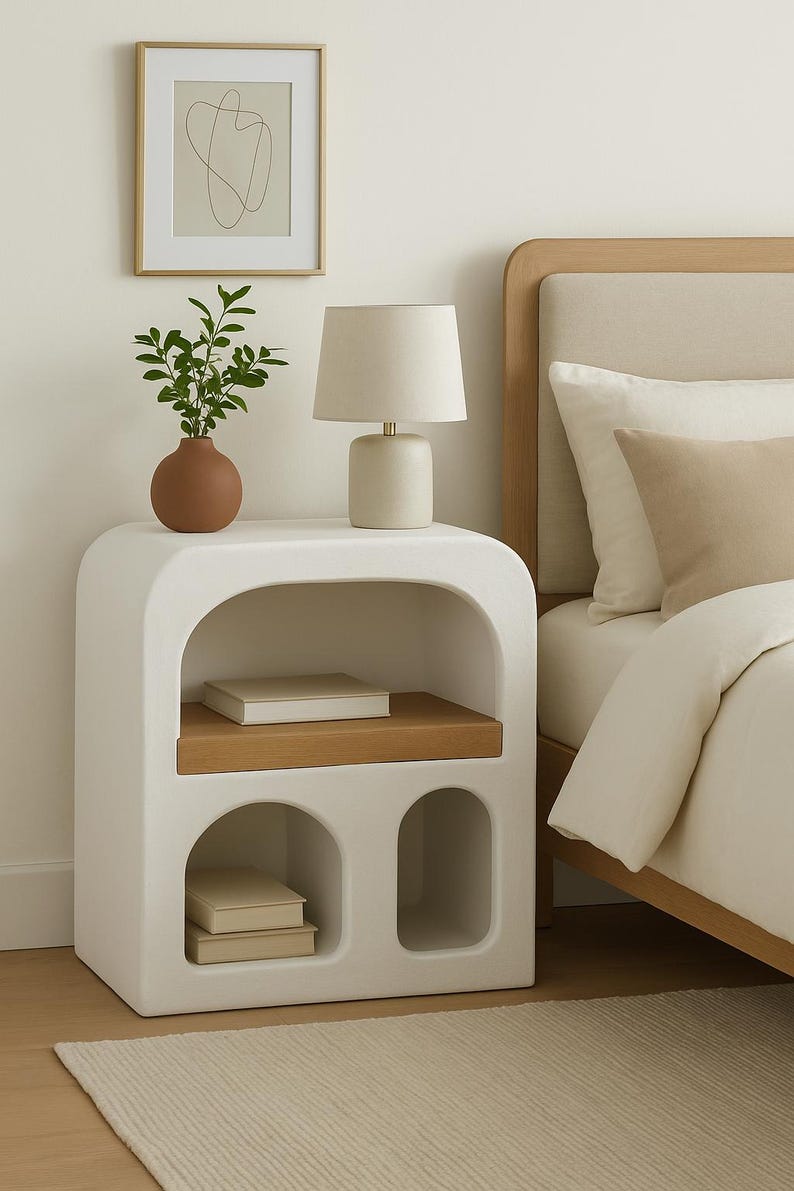 Puede incluir: Una mesita de noche blanca moderna con un dise&ntilde;o curvo, con una estanter&iacute;a de madera y recortes arqueados. Una l&aacute;mpara beige, un peque&ntilde;o jarr&oacute;n de terracota con vegetaci&oacute;n y libros est&aacute;n colocados sobre la mesa. Una obra de arte abstracta enmarcada cuelga en la pared.
