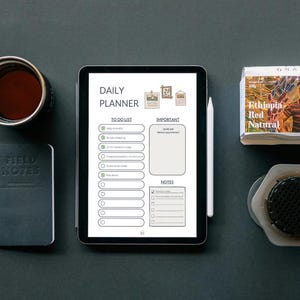 Könnte beinhalten: Eine Draufsicht mit einem digitalen Tablet, das einen Tagesplaner, eine Kaffeetasse, ein Notizbuch, einen Stift, eine Packung Kaffeebohnen und eine Kaffeemaschine zeigt. Das Tablet hat einen weißen Stift. Die Gegenstände liegen auf einer dunklen Oberfläche.