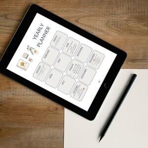 Könnte beinhalten: Ein schwarzes Tablet mit einem Jahresplaner mit Monaten und Daten, einem schwarzen Bleistift und einem leeren weißen Blatt Papier auf einer Holzoberfläche. Der Planer hat ein minimalistisches Design mit hellgrauem Hintergrund und weißem Text. Der Text "YEARLY PLANNER" steht oben.
