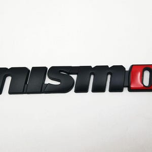 Puede incluir: Emblema negro "NISMO" con un acento "O" rojo y negro. El emblema es un accesorio para automóvil, probablemente para el exterior de un vehículo. Las letras son en negrita y estilizadas, con un acabado brillante en la sección roja.