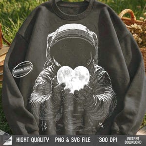 May include: Dark gray sweatshirt featuring a black and white graphic of an astronaut holding a glowing heart-shaped object. The design includes the text "ORIGINAL DESIGN" on a white oval. The sweatshirt has a crew neck and long sleeves.