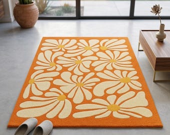 May include: Orange rectangular rug with a large daisy pattern in cream and yellow. The rug is placed on a concrete floor, with a pair of gray slippers in the foreground. The rug's design is reminiscent of the 1970s.
