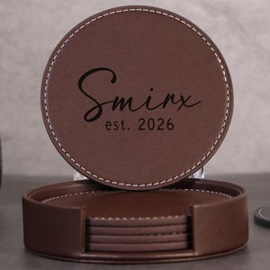 Op de afbeelding: Een set bruine leren onderzetters met witte stiksels. Eén onderzetter wordt bovenop de set weergegeven, met de naam "Sminx" en het jaar "est. 2026" in een stijlvol lettertype. De onderzetters zijn rond.