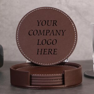 Könnte beinhalten: Ein Set aus runden, braunen Lederuntersetzern mit weißen Nähten, die in einem passenden runden Halter aufbewahrt werden. Der oberste Untersetzer trägt den eingravierten Text "YOUR COMPANY LOGO HERE" in einer dunklen Serifenschrift.