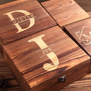 May include: Four wooden boxes with engraved initials and names. The boxes are made of dark brown wood with a natural grain. The letters and names are in a lighter color, creating a contrast. One box reads "DIETER", another "JYE".