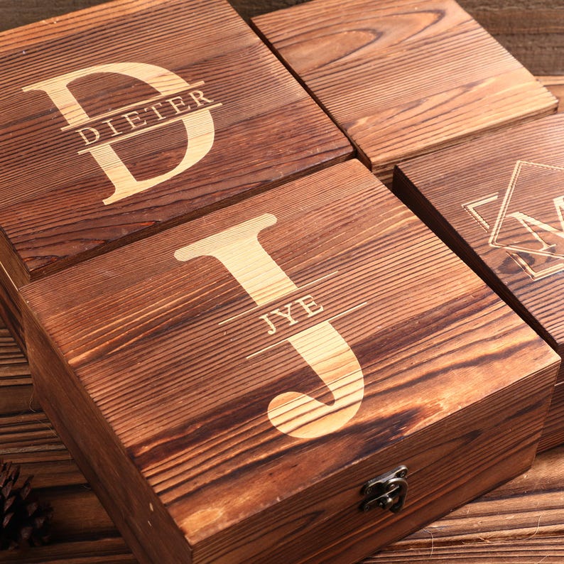 May include: Four wooden boxes with engraved initials and names. The boxes are made of dark brown wood with a natural grain. The letters and names are in a lighter color, creating a contrast. One box reads "DIETER", another "JYE".