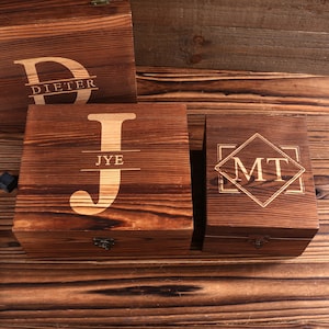May include: A collection of wooden boxes with engraved initials and names. The boxes are various sizes and have a dark brown finish. One box has the letter "J" with the name "JYE" below it. Another box has the initials "MT".