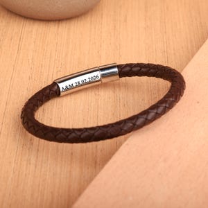 May include: A brown braided leather bracelet with a silver clasp. The clasp is engraved with the initials "A&M" and the date "28.02.2026". The bracelet is a circular shape and is displayed on a light brown wooden surface.