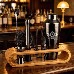 May include: A black cocktail shaker set with a bamboo stand. The shaker features a white crest logo with the text RUSSELL. The set includes a long twisted bar spoon, a jigger, a muddler, a strainer, a pourer, and a corkscrew, all in black metal.