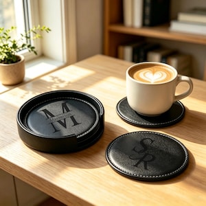 Puede incluir: Un juego de posavasos redondos de cuero negro con costuras blancas y monogramas personalizados. Un posavasos está en un soporte negro a juego, otro bajo una taza de cerámica blanca con arte latte y uno más muestra las letras SR grabadas.