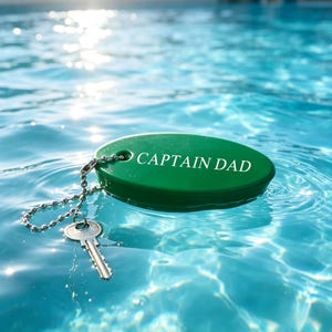 Könnte beinhalten: Ein grüner, ovaler, schwimmfähiger Schlüsselanhänger mit dem weißen Schriftzug CAPTAIN DAD auf der Oberfläche. Eine silberne Metallkugelkette ist am Anhänger befestigt und mit einem einzelnen silbernen Metallschlüssel verbunden, der auf dem Wasser liegt.