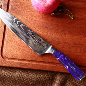 May include: A stainless steel chef's knife with a patterned blade and a purple and white marbled handle. The blade is engraved with the text "10 YEARS OF US" and a date. The knife rests on a wooden cutting board with a red apple and cherries.