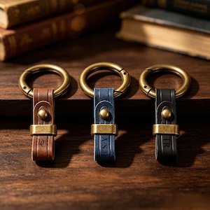 Op de afbeelding: Drie leren sleutelhangers met messingkleurige hardware. De sleutelhangers zijn bruin, blauw en zwart. Elke sleutelhanger heeft een metalen ring en een leren bandje met gegraveerde initialen. Ze worden gepresenteerd op een houten oppervlak.