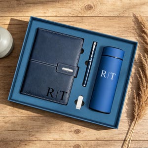 Peut inclure: Un coffret cadeau bleu marine dans une boîte rectangulaire comprenant un carnet à texture cuir avec les lettres R|T, un stylo en métal bleu marine assorti, une petite clé USB argentée et une gourde cylindrique bleu marine avec R|T.