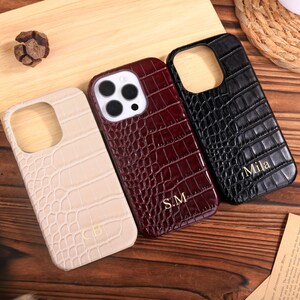 Puede incluir: Tres fundas para teléfono con textura de piel de cocodrilo se muestran sobre una superficie de madera. Las fundas son de color crema, burdeos y negro. La funda burdeos tiene las iniciales "S.M." en dorado, y la funda negra tiene el nombre "Mila" en dorado.