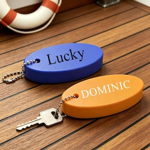 May include: Two oval-shaped floating keychains, one blue with the word Lucky printed in black and one orange with the word DOMINIC printed in white. Each keychain features a silver-toned metal ball chain and a silver-toned metal key attached.