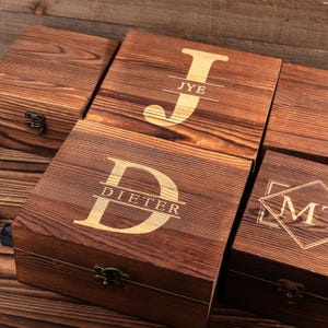 May include: Wooden boxes with engraved initials and names. The boxes are made of dark brown wood and feature gold-colored lettering. The initials "JYE", "D", and "M" are visible, along with the name "DIETER". Each box has a metal clasp.