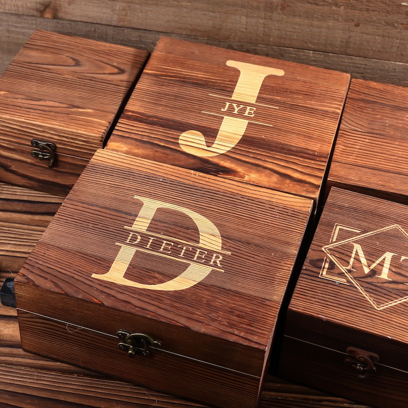May include: Wooden boxes with engraved initials and names. The boxes are made of dark brown wood and feature gold-colored lettering. The initials "JYE", "D", and "M" are visible, along with the name "DIETER". Each box has a metal clasp.