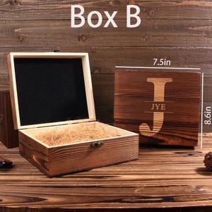 May include: Wooden gift box with an open lid, revealing a black interior and straw-like filler. A closed wooden box with the letter "J" and the initials "JYE" is next to it. The closed box measures 7.5 inches by 8.6 inches.