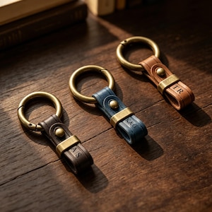 May include: Three leather keyrings with brass fittings are displayed on a wooden surface. The keyrings are brown, blue, and tan. Each keyring has initials engraved on the leather strap. The keyrings have a large brass ring.
