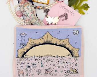 Puede incluir: Una bolsa de tela rosa con cierre de cremallera presenta una ilustración caprichosa de un paisaje desértico con un gran sol, cactus y varios animales. La bolsa está llena de una variedad de artículos, incluyendo un collar de perlas, una pequeña lata de caramelos de jengibre, una tira de cabina de fotos vintage, una pequeña muñeca, un juego de dientes de plástico y una tarjeta con el texto "Madonna Inn".