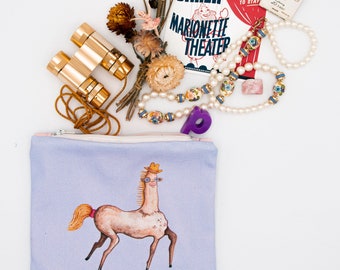 Puede incluir: Una bolsa de cremallera lavanda con una ilustración caprichosa de un caballo que lleva un sombrero de vaquero y gafas. La bolsa está rodeada de varios artículos, incluyendo un par de binoculares de oro, una cadena de perlas, un programa de teatro vintage y un pequeño caballo de madera.