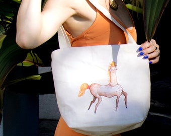 Puede incluir: Una bolsa de lona blanca con un gráfico de un caballo con un sombrero y gafas de sol. El caballo tiene un cuello largo y está de pie sobre sus patas traseras.