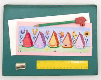 Puede incluir: Una tarjeta rosa y blanca con una ilustración colorida de cinco casas con caras. Las casas están alineadas en una fila y tienen techos y puertas de diferentes colores. Las casas están rodeadas de flores y plantas. La tarjeta está sobre una superficie verde con un lápiz, una goma de borrar y una regla.