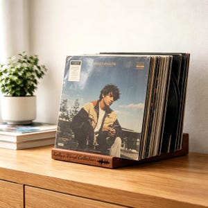 Pode incluir: Um suporte de madeira para discos de vinil exibindo uma coleção de discos. A capa do disco frontal apresenta um homem de jaqueta. O suporte é gravado com "Kelly's Vinyl Collection" e um design de onda sonora.