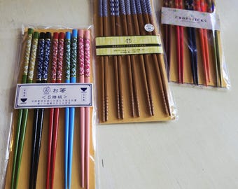 May include: Three sets of five pairs of chopsticks in clear plastic packaging. One set features colorful floral patterns in green, black, red, blue, and pink. Another set has brown bamboo sticks with blue patterned tops. The third set has dark tones.
