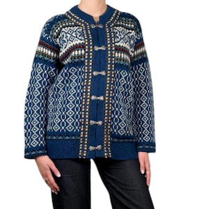Può includere: Un cardigan blu con un motivo geometrico e chiusure a toggle. Il maglione presenta accenti bianchi, rossi e beige. La persona indossa jeans di denim scuro e scarpe nere. Il cardigan è uno stile classico.