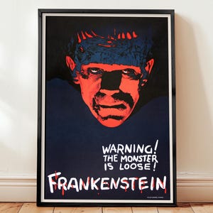 May include: Framed poster featuring a red and blue image of Frankenstein's monster. The poster has the text "WARNING! THE MONSTER IS LOOSE! FRANKENSTEIN" in white and red lettering. The poster is in a black frame and is displayed on a white wall.