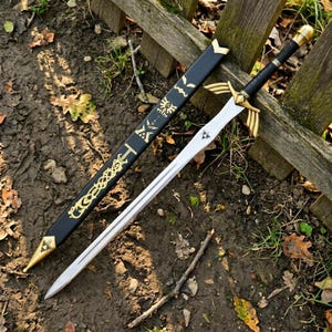Peut inclure: Une &eacute;p&eacute;e en m&eacute;tal avec une lame argent&eacute;e, une garde dor&eacute;e aux extensions en forme d'ailes et un manche noir avec un pommeau dor&eacute;. &Agrave; c&ocirc;t&eacute; se trouve un fourreau noir orn&eacute; de symboles dor&eacute;s, dont un embl&egrave;me en forme de triforce.