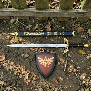 Peut inclure: Une &eacute;p&eacute;e argent&eacute;e avec une poign&eacute;e noire et une garde dor&eacute;e, un fourreau noir orn&eacute; de motifs d&eacute;coratifs dor&eacute;s, et un bouclier au centre marron avec un embl&egrave;me dor&eacute; repr&eacute;sentant un oiseau et un triangle.